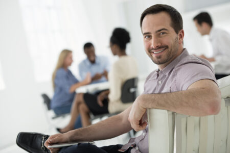 A fresh white building interior, flooded with light. A group of people meeting. One person looking over his shoulder and away from the group. Holding a digital tablet.